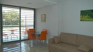 Un salon moderne avec un canapé beige, une table basse en verre et des fauteuils orange. On y trouve une grande fenêtre avec des stores, un tableau encadré au mur et une peinture représentant un chemin bordé d’arbres.