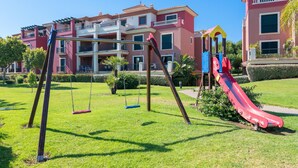 A playground with swings and a slide, surrounded by well-maintained grass and a building with balconies.