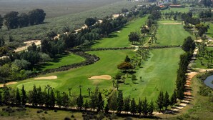 A golf course with sand bunkers, water bodies, and a cityscape in the background.