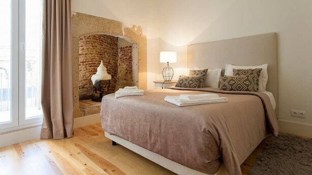 Un dormitorio con pared de ladrillo, cama con colcha color arena, mesita de noche con lámpara y ventana con cortinas.