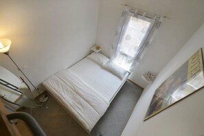 A small, single-bed room with a bed, a nightstand, a lamp, and a window with a curtain.