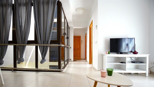 Un salón moderno con un separador de vidrio, un televisor sobre un soporte blanco y una pequeña mesa con una taza verde.