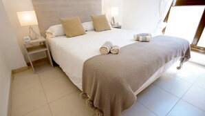 Una cama bien hecha con una manta tejida y toallas enrolladas, una mesita de noche con una lámpara y un buró con un cargador de teléfono.
