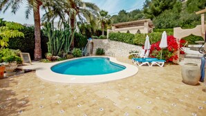 Ein Swimmingpool mit Steinmauer, Palmen und einer Sonnenterrasse mit Liegestühlen und Sonnenschirmen.