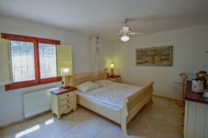 A bedroom with a bed, bedside tables, a ceiling fan, a window with curtains, and a painting on the wall. - Chrisuli - well furnished villa with panoramic views in Moraira (Benimeit)