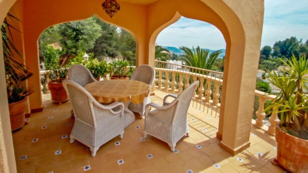 Eine überdachte Terrasse mit einem runden Tisch und vier Korbstühlen, umgeben von Topfpflanzen und einem Fliesenboden.