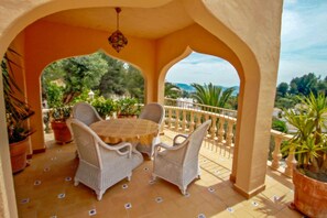 Une terrasse couverte avec une table ronde et quatre chaises en osier, entourée de plantes en pot et dotée d’un sol carrelé.