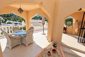 A covered outdoor seating area with wicker furniture and a glass table, surrounded by a white balustrade, overlooking a residential area. - Chrisuli - well furnished villa with panoramic views in Moraira (Benimeit)