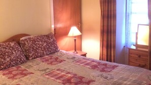 A bedroom with a wooden ceiling, a bed with floral and checkered bedding, a nightstand with a lamp, and a window with curtains. - Pond Cottage, Carmichael Country Cottages. Pets welcome. (Biggar)