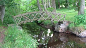 A wooden bridge over a calm stream in a lush green forest. - Pond Cottage, Carmichael Country Cottages. Pets welcome. (Biggar)