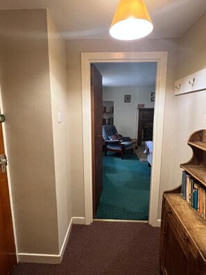 A hallway leading to a living room with a green carpet, a blue armchair, and a wooden cabinet with books. - Pond Cottage, Carmichael Country Cottages. Pets welcome. (Biggar)