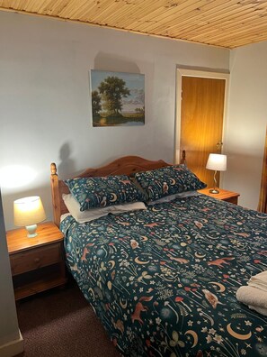 A bedroom with a wooden headboard, a bed with a patterned duvet, a nightstand with a lamp, and a painting on the wall. - Pond Cottage, Carmichael Country Cottages. Pets welcome. (Biggar)