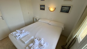 Una habitación de hotel con una cama, dos toallas dispuestas en forma de flores, una mesita de noche, una lámpara de pared y cuadros en la pared.
