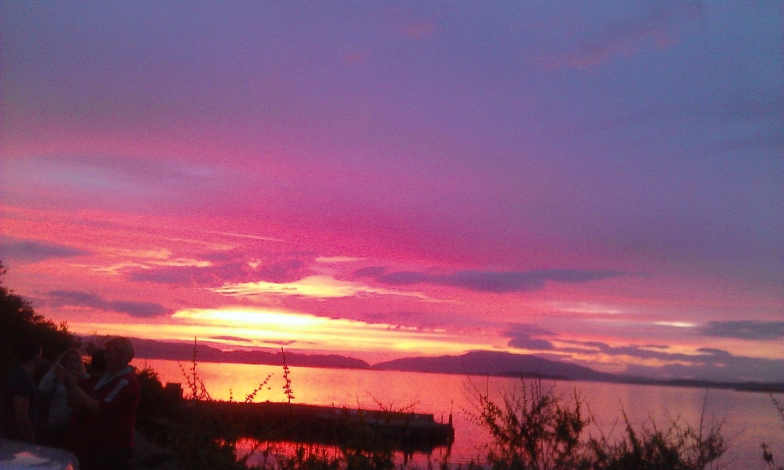 Ein Sonnenuntergang über einem Gewässer mit einem Steg und Vegetation im Vordergrund.