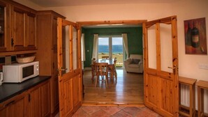 A kitchen with wooden cabinets, a microwave, and a dining area with a table and chairs. There is a view of the beach through a glass door. - Cottage 259 - Renvyle - sleeps 6 guests  in 3 bedrooms (Renvyle)