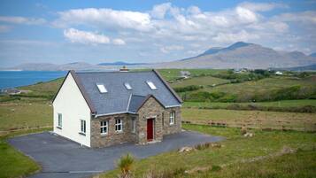 A stone house with a red door, a sloped roof, and a view of the sea and hills in the background.