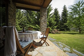 An outdoor patio with a stone table and wooden chairs, surrounded by a stone wall and lush greenery.