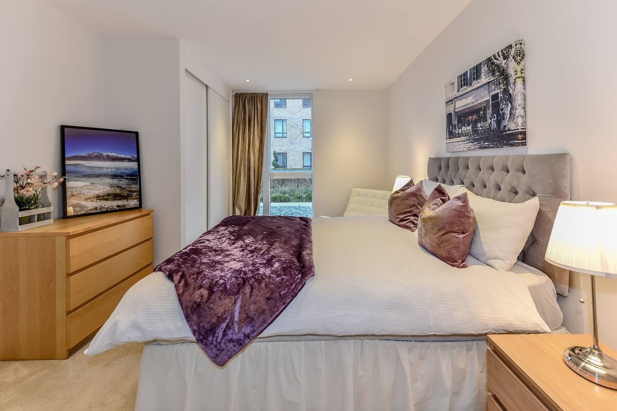 A modern bedroom with a large bed, a nightstand, a dresser, and a window with curtains.
