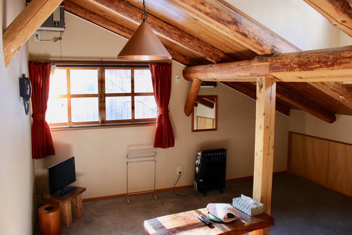 A room with a sloped ceiling, wooden beams, a window with red curtains, a radiator, a television on a small wooden stand, and a table with a remote control and tissues.