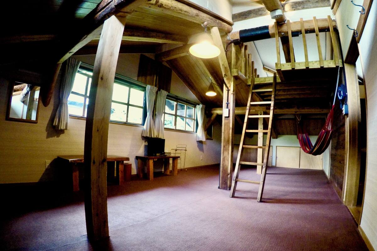 A rustic room with exposed wooden beams, a loft bed, a ladder, and a hammock.