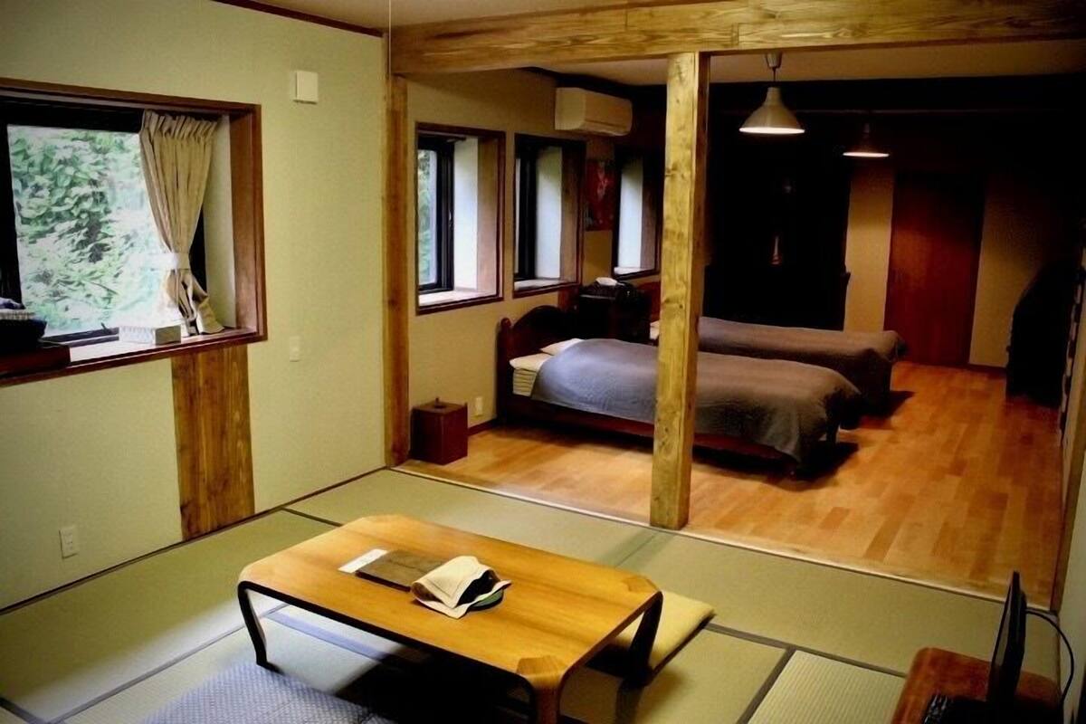 A traditional Japanese-style room with tatami flooring, a wooden table, a bed with a futon, and a view of greenery outside.