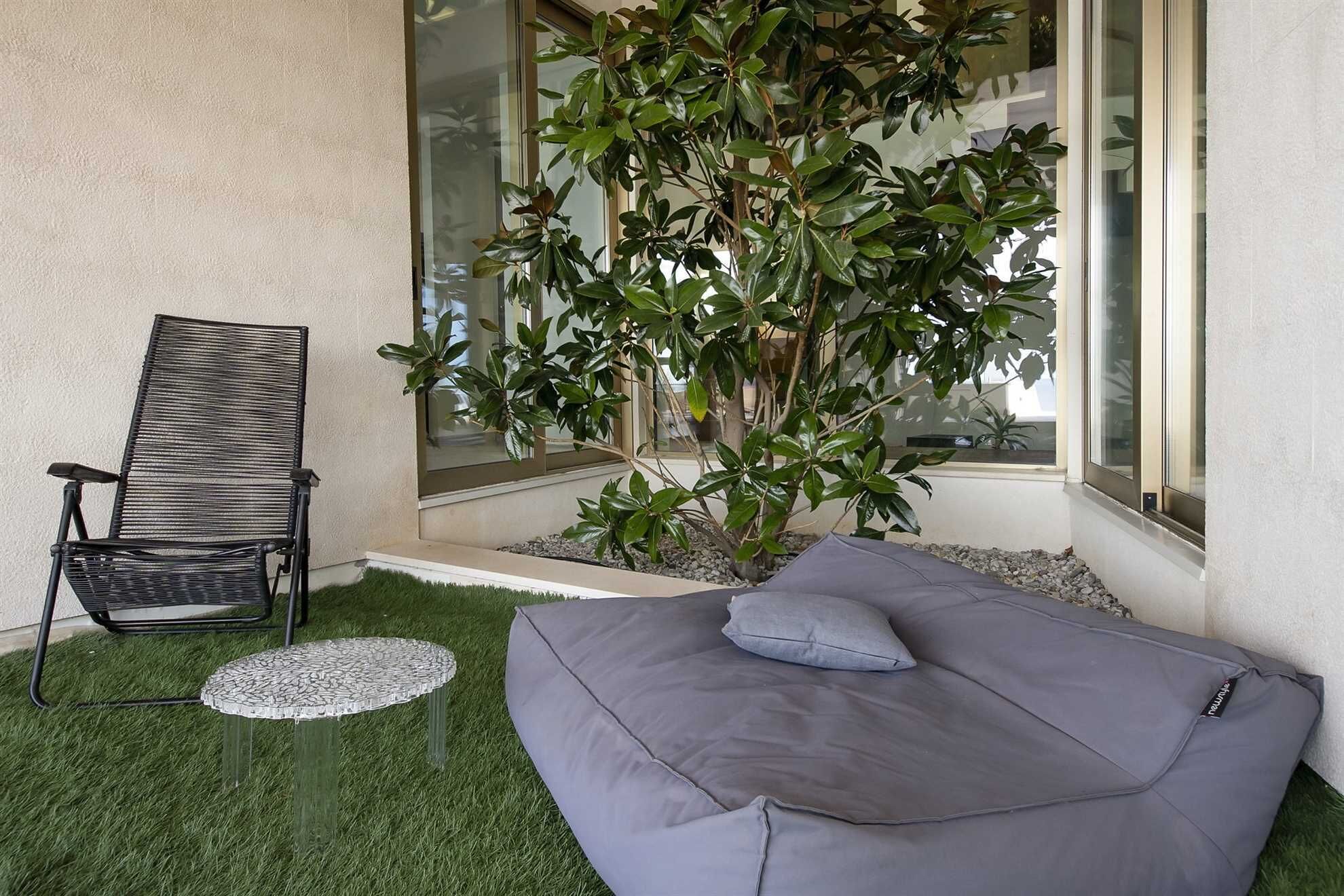 A modern outdoor seating area with a cushioned bench and a metal chair, surrounded by a small tree and a glass window.