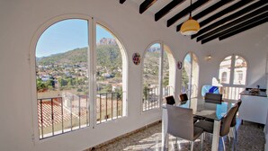 Zona de menjador amb taula i cadires, grans finestres d'arc amb vista a una població i muntanyes, i zona de cuina amb lluminària penjant.