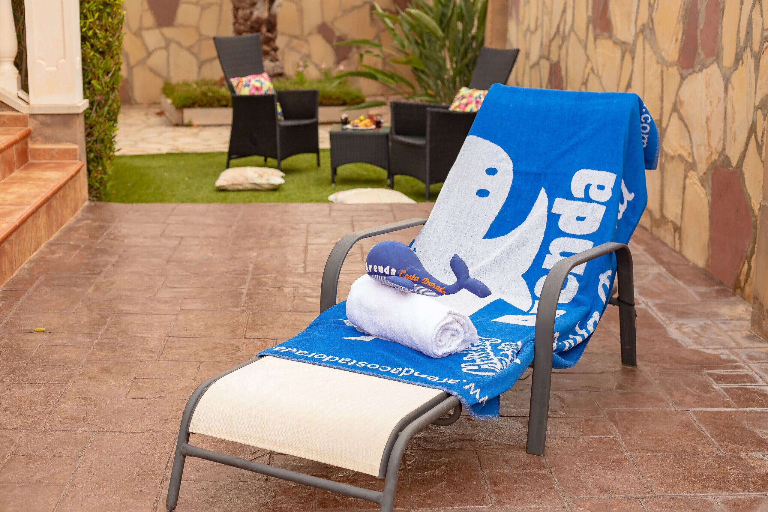 Una silla de descanso con una toalla azul y otra enrollada encima, sobre un patio con baldosas, una pared de piedra y muebles de exterior al fondo.