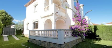 A white building with a stone base, a balcony, and a garden with purple flowers.