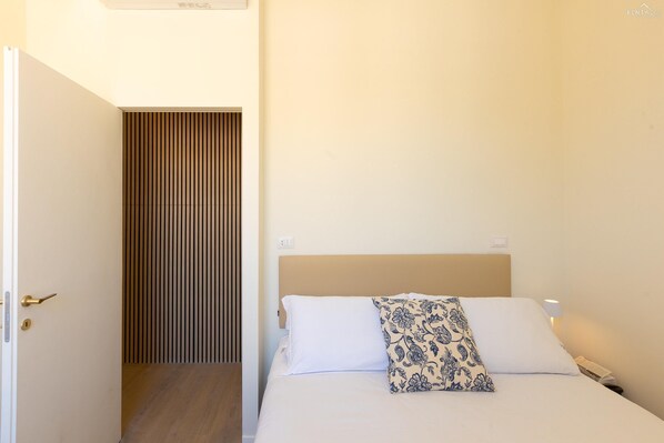A hotel room with a bed, a bedside table with a lamp, and a door leading to a corridor.
