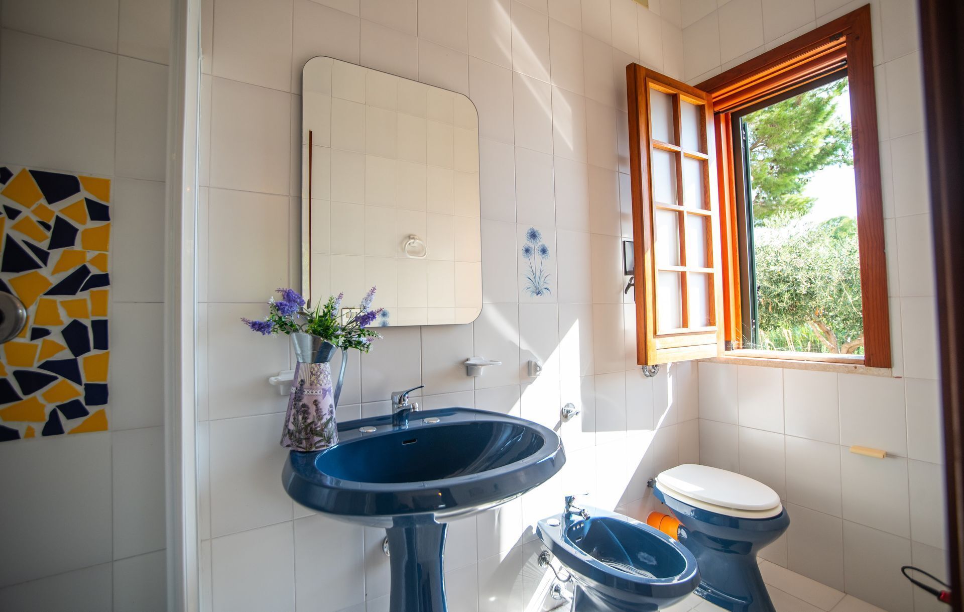 A bathroom with a blue sink, a toilet, a mirror, and a window with an open shutter.