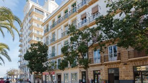 A multi-story building with balconies, storefronts, and a pedestrian area with trees.