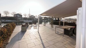 Un patio techado al aire libre con suelo de baldosas, arreglos de asientos y vistas a un canal con barcos.