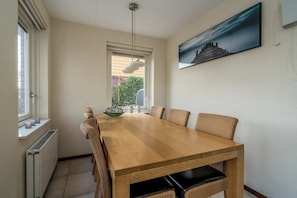 Un comedor con una mesa de madera, sillas de mimbre, una lámpara colgante, una ventana con persianas y un cuadro en la pared.