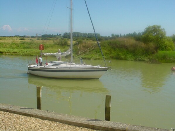 Un voilier est amarré sur une rivière paisible, bordée d’une rive herbeuse, avec des arbres en arrière-plan.