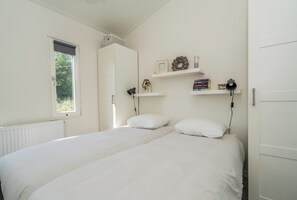Un dormitorio con cama, ventana, radiador, armario y estante con libros y objetos decorativos.