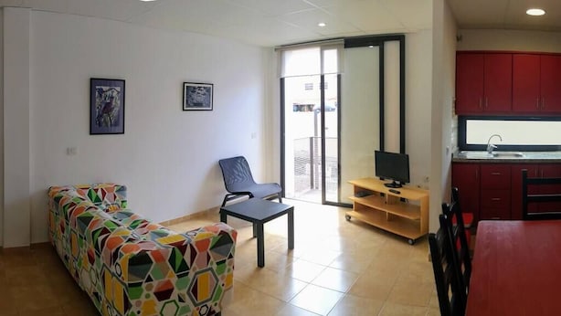 Una sala de estar con un sofá colorido, una silla, una mesita y un televisor. Se observa una zona de cocina con armarios rojos y un lavamanos.