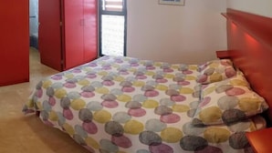 Un dormitorio con una cama, un armario rojo, una puerta que da a otra habitación y una ventana con persianas.