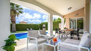 Un espace extérieur abrité comprenant une piscine, des chaises longues, une table avec des boissons et une vue sur une maison et un palmier.