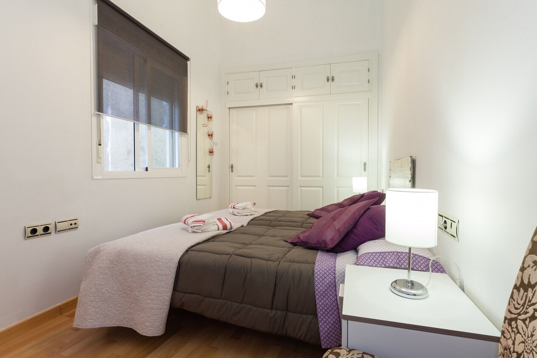 Quarto com cama, mesa de cabeceira com abajur, janela com persianas e guarda-roupa.