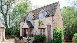 A stone house with a pitched roof, a small porch with a table and chairs, and a brick wall with a window.
