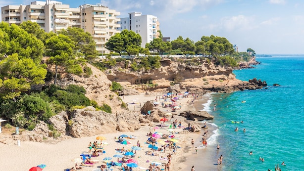 Ein Strand mit badenden und sonnenden Menschen, umgeben von Gebäuden und Bäumen.