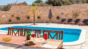 A poolside dining area with a stone wall, a table set for two with drinks and a bottle, and lounge chairs along the pool. - Villa Anfiteatro Adriatico with Pool (Murter)