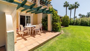 A patio with a table and chairs, a pergola, and a well-maintained lawn with palm trees. - Hoyo I - Isla Canela Golf GHHI15B2P65 (Isla Canela)