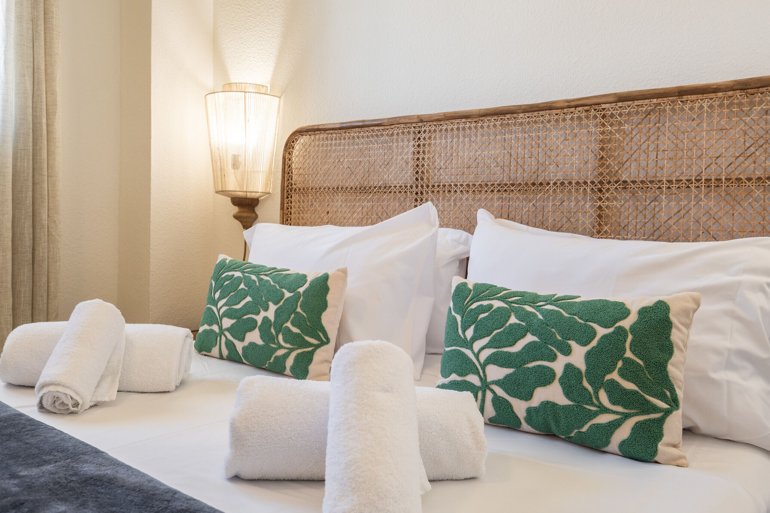 Una cama bien hecha con sábanas blancas y almohadas estampadas con motivos de hojas, un cabecero tejido y una lámpara de noche.