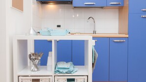 Une cuisine avec des meubles bleus, un plan de travail blanc et une étagère blanche où sont rangés des ustensiles et des torchons.