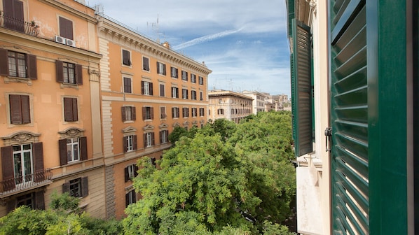 A row of historic buildings with uniform window shutters and balconies, surrounded by lush greenery.