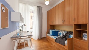 Une petite pièce avec un lit en bois, un bureau avec une lampe, un tableau d’affichage en liège et une fenêtre avec des rideaux.