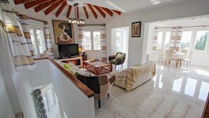 A spacious living area with a sofa, a coffee table, and a TV. There are curtains, a painting, and a dining area with chairs and a table. - Gila - sea view villa with private pool in Benissa (Benissa)