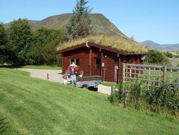 A rustic wooden cabin with a thatched roof, a gravel path leading to it, and a wooden fence in the foreground.